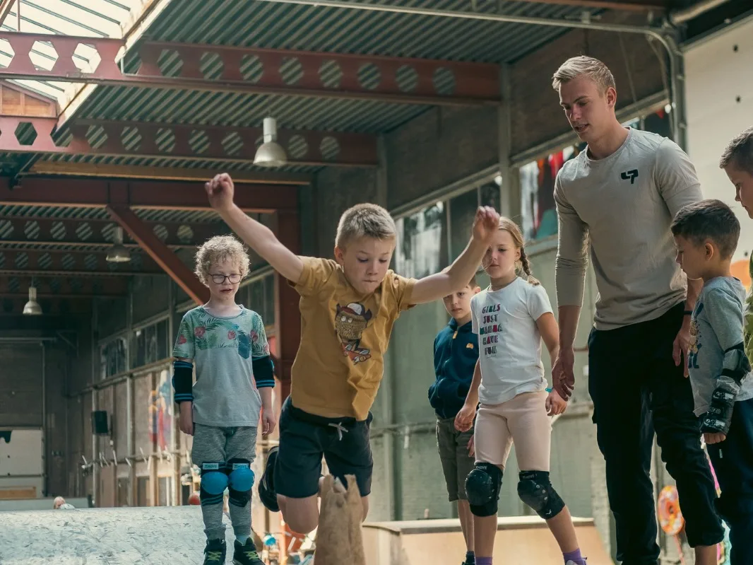 Leerling springt met gespreide armen over een pion tijdens een DAREcademy-freerunles, de groep en de coach kijken toe